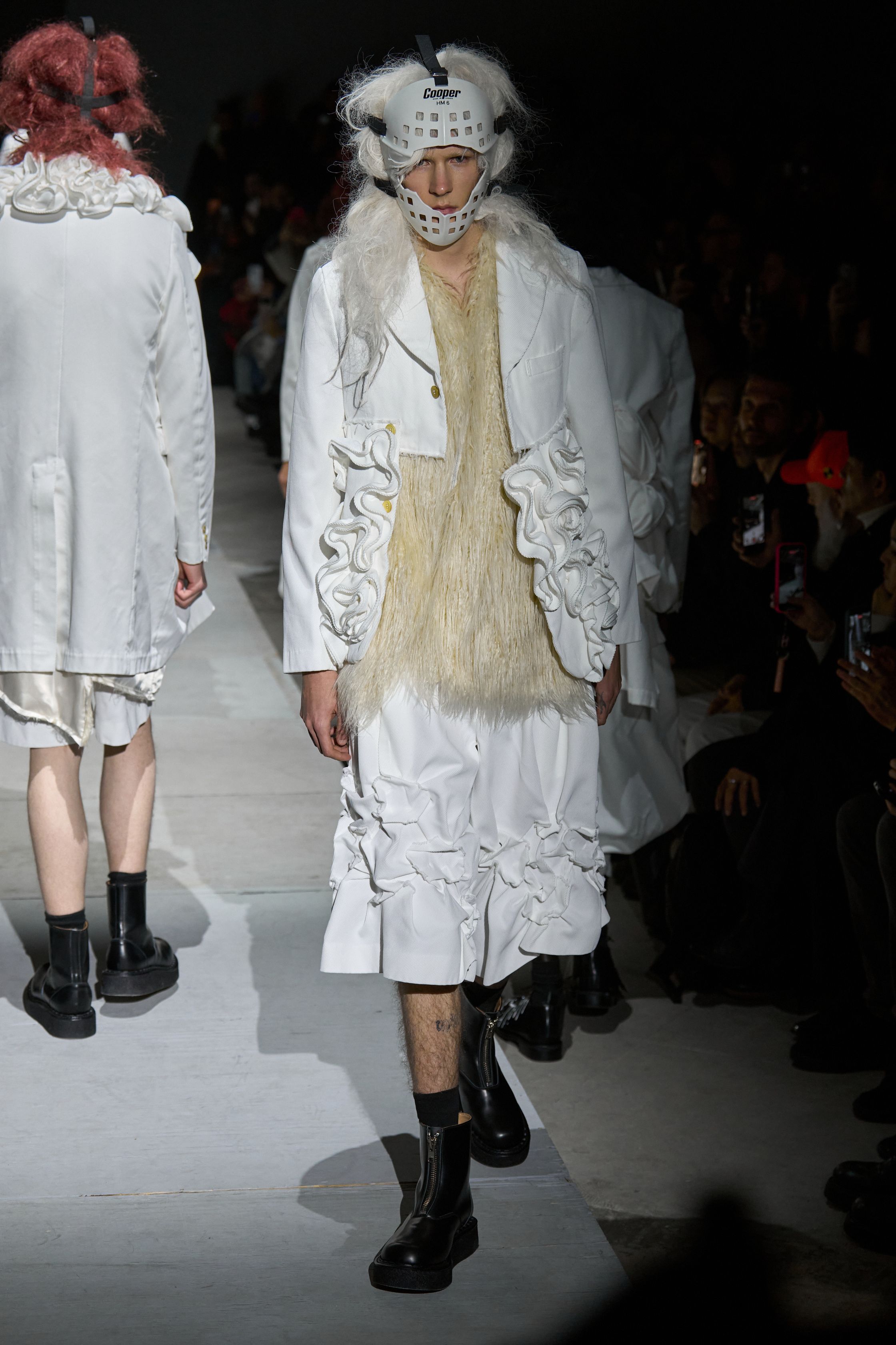 Comme des Garçons Homme Plus 2026秋冬以极端造型继续挑战“男性身体”的社会想象。面具、假发、解构西装与异化比例营造出近乎仪式化的压迫氛围，黑白灰成为思想载体而非装饰。这是一场关于权力、恐惧与集体意识的时装实验，服装不再服务于穿着，而是成为观念的武器-顽石频道趋势研究